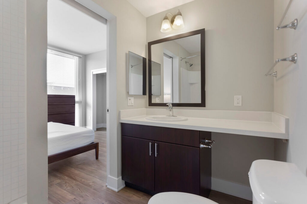 Modern bathroom with a dark wood vanity, large mirror, single sink, and toilet. The bathroom opens to a bedroom with a bed, wooden floor, and large window letting in natural light.