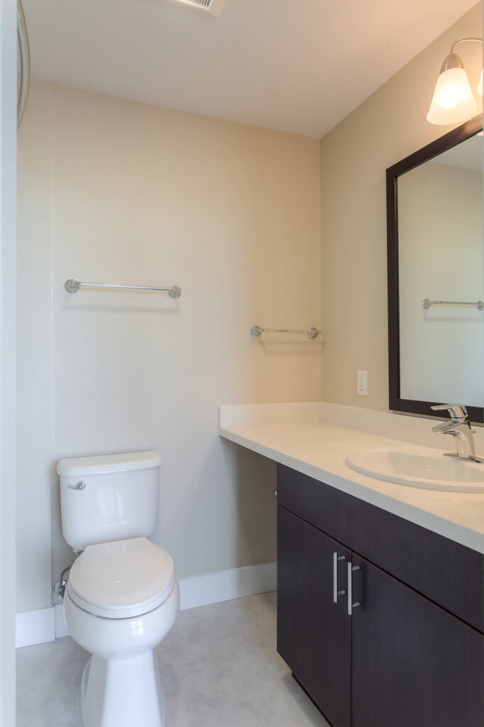A simple bathroom with a toilet, a dark wood vanity, a sink, a large mirror with a light above it, and empty towel bars on beige walls. The flooring is light-colored.