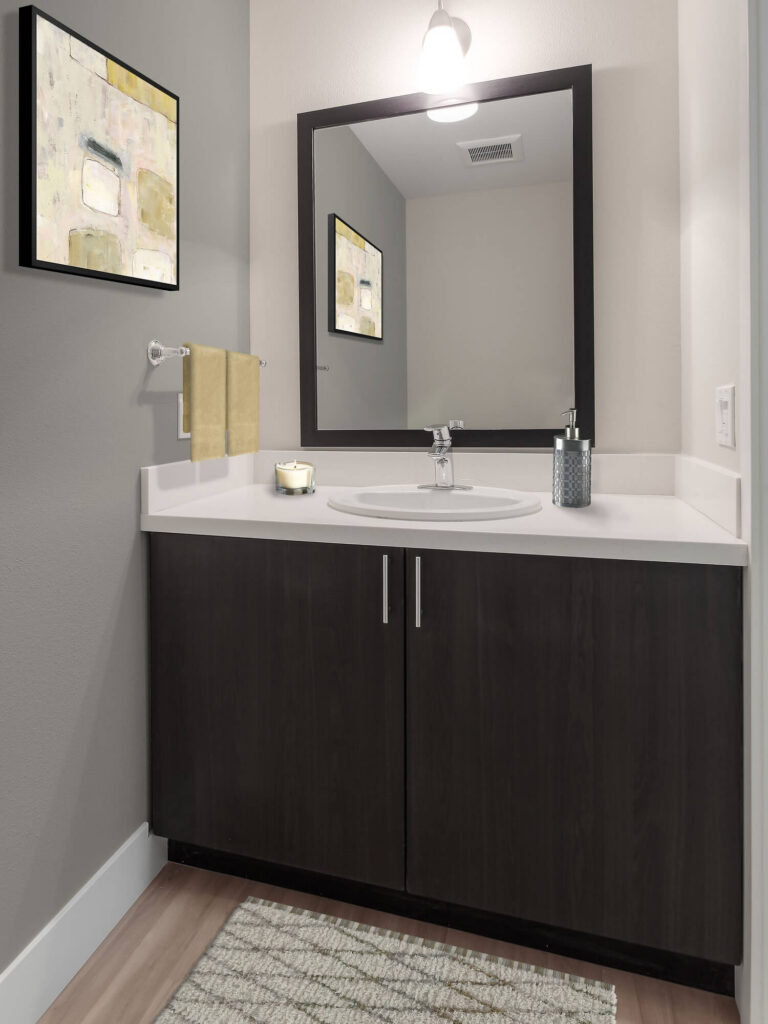 Modern bathroom with a dark wood vanity, white countertop, large mirror, light fixture above, yellow towels, wall art, and a striped rug on a wooden floor. Soap dispenser and toiletries are on the counter.