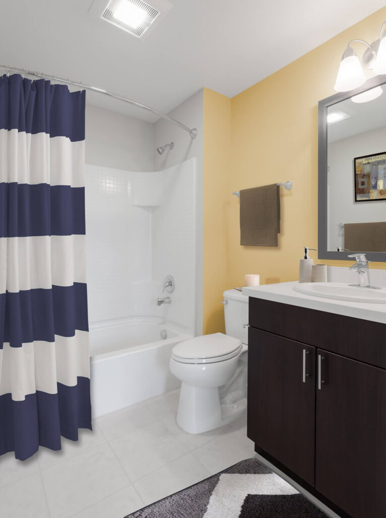 Modern bathroom with a white tub and toilet, navy-and-white striped shower curtain, yellow wall, dark brown vanity, rectangular mirror, towel, soap dispenser, and patterned rug.