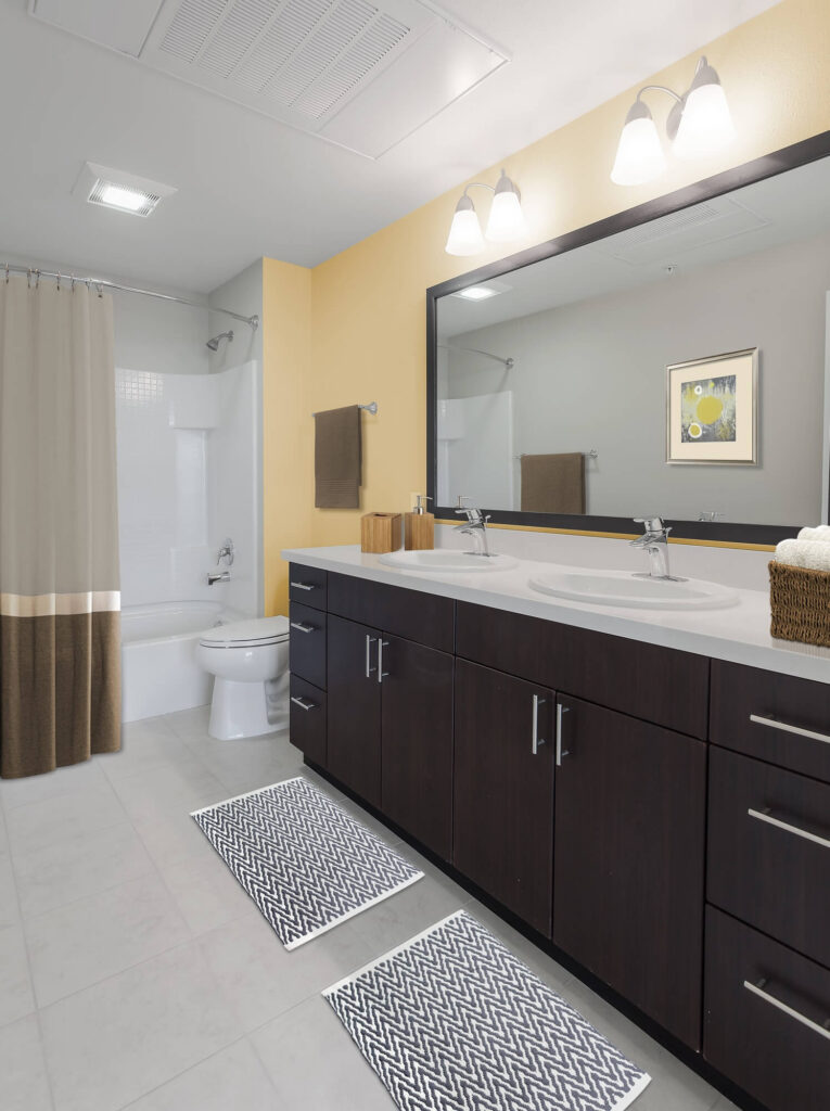 Modern bathroom with double sink vanity, large mirror, two chevron-patterned rugs, shower with curtain, toilet, towel rack, and yellow accent wall with a framed picture. Bright lighting overhead.