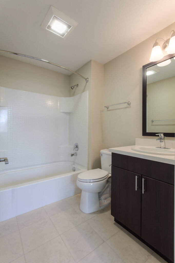 Modern bathroom with a white bathtub and shower, toilet, dark wood vanity with sink, large mirror, and wall light fixture. The floor is tiled in light beige.