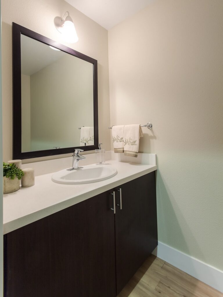 Modern bathroom vanity with a white countertop, dark brown cabinets, a rectangular mirror, single faucet sink, towels on a rack, and a small plant decoration on the left side.