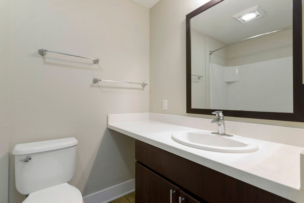 Modern bathroom with a white toilet, dark wood vanity, white countertop, sink with chrome faucet, large mirror, and empty towel racks on the beige wall. Shower area with a curved rod is visible in the background.