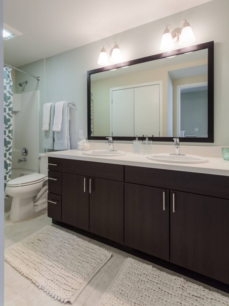 Modern bathroom with double sinks, dark wood cabinets, large mirror, two light fixtures, white rugs, and a toilet next to a tub with a blue and white shower curtain. Towels hang on a rack near the shower.