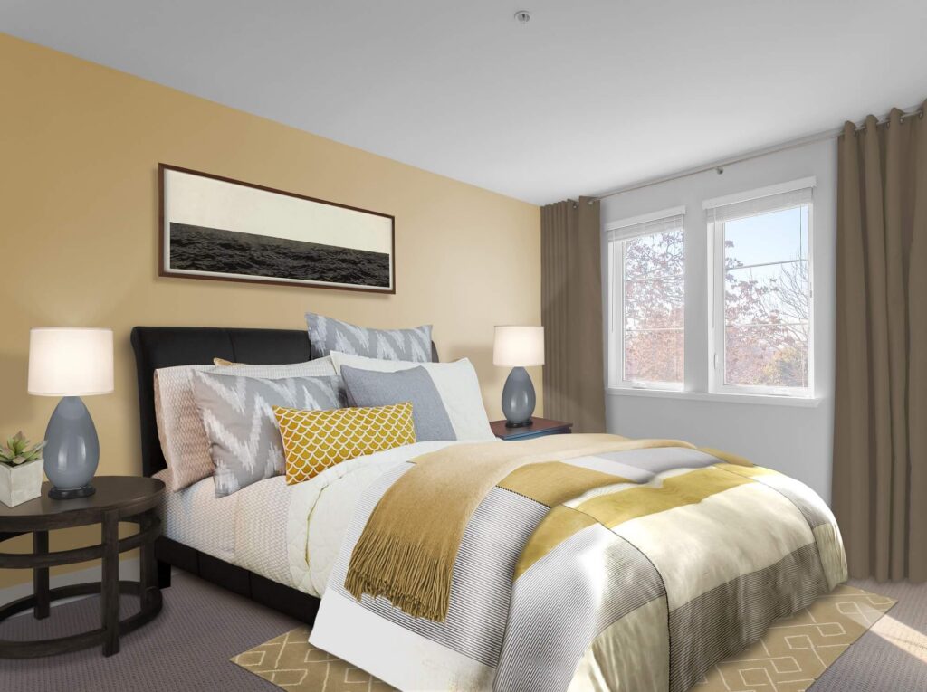 A cozy bedroom with a neatly made bed, yellow and gray bedding, two bedside tables with lamps, beige walls, and two windows with beige curtains letting in natural light.