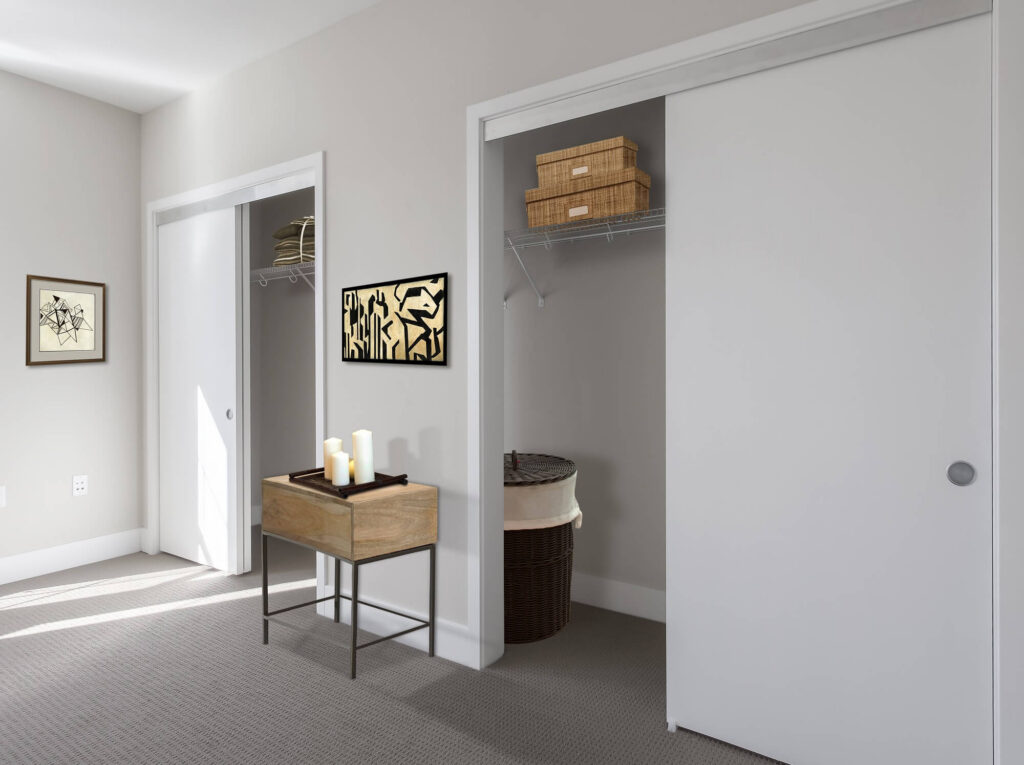 A bedroom with two closets, one open showing shelves with baskets and a laundry hamper. A small table with candles stands against the wall, and framed art hangs above it. The room has neutral colors and carpeted flooring.