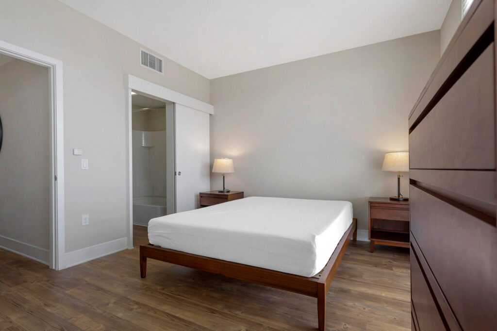 A modern, minimal bedroom with a wooden bed frame, white bedding, two bedside tables with lamps, a dresser, wooden floor, and an open door leading to a bathroom. Walls are painted light gray.