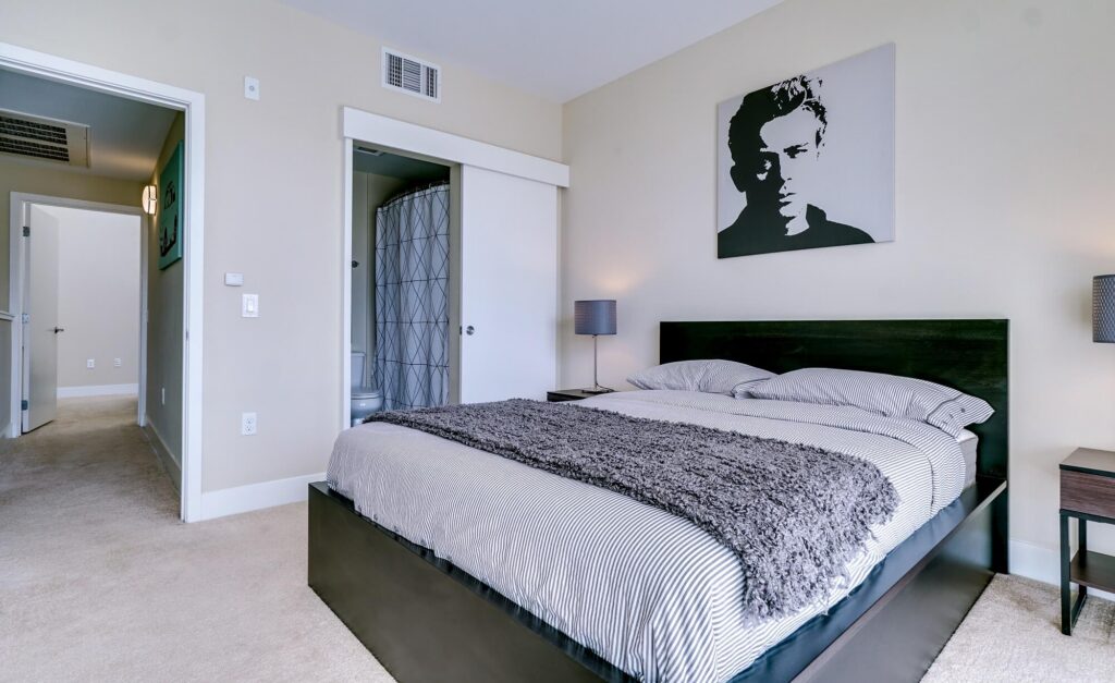 A modern bedroom with a black bed frame, gray bedding, two nightstands with lamps, and wall art above the bed. An open door reveals a bathroom with a shower curtain, and a hallway leads to another room.