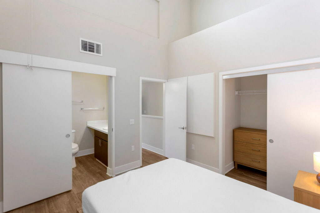 A bright bedroom with a bed, open closets, wooden floors, a small dresser, and a sliding door revealing a bathroom with a sink and toilet. A lamp sits on a nightstand beside the bed.