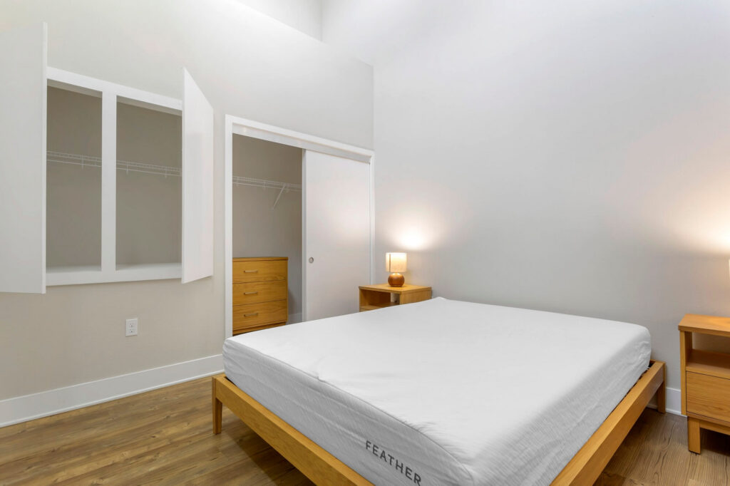 Minimalist bedroom with a wooden bed frame, white mattress, two wooden nightstands with lamps, a wooden dresser, and open closet doors. Walls are white, and the floor is light wood.