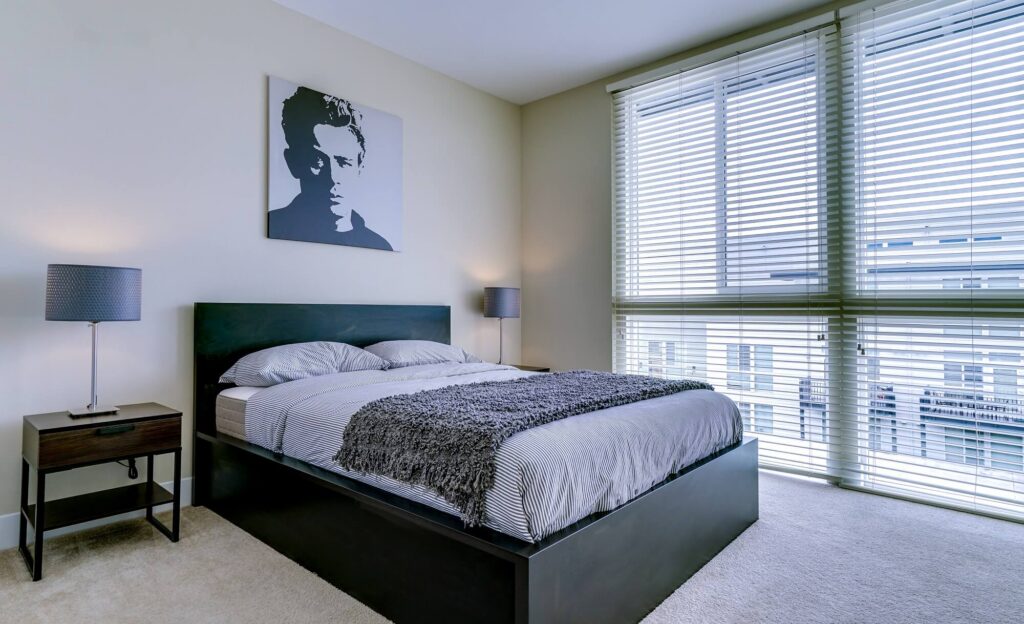 Modern bedroom with a black bed, gray bedding, two nightstands with lamps, large window with blinds, and a monochrome portrait above the bed. Natural light fills the room.