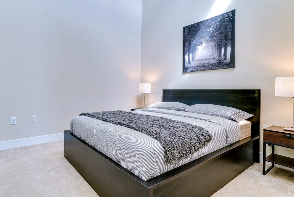 Modern bedroom with a dark wood bed, striped bedding, gray throw blanket, two bedside tables with lamps, and a black-and-white forest photo on the wall.