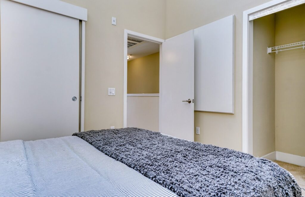 A bedroom with a bed in the foreground, a gray blanket, an open closet with a wire shelf, a closed sliding door, and an open door leading to a hallway.