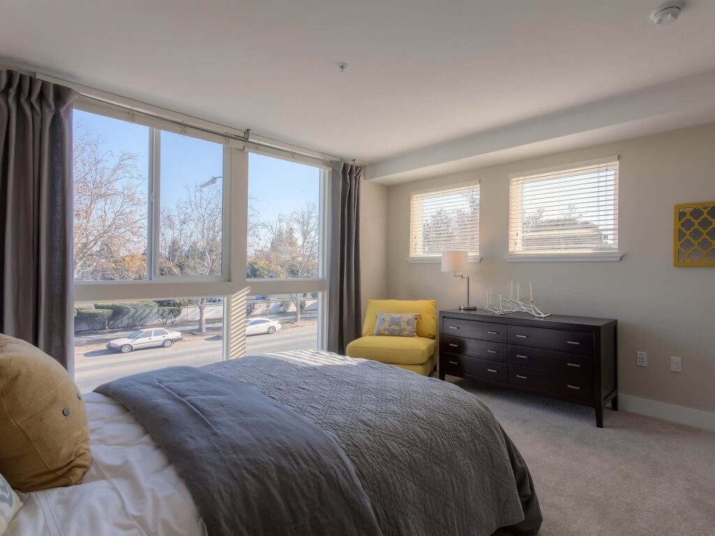 Sunny bedroom with large windows, gray bedding, a yellow chair, dark dresser, and light walls. Outside, cars are parked along the street lined with trees.