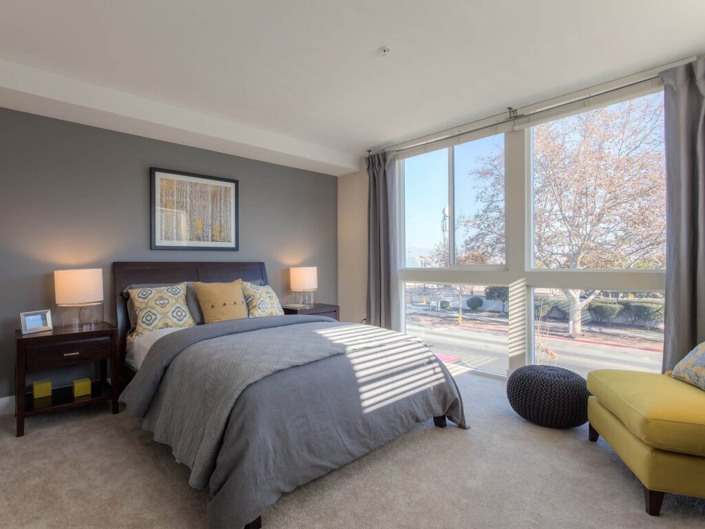 Modern bedroom with a large window, gray walls, a neatly made bed with gray bedding, two bedside tables with lamps, a yellow armchair, a black pouf, and trees visible outside.