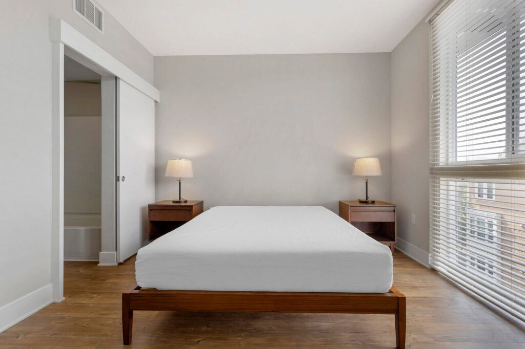 Minimalist bedroom with a wooden bed frame, white bedding, two bedside tables with lamps, light wood floor, and large window with blinds letting in natural light.
