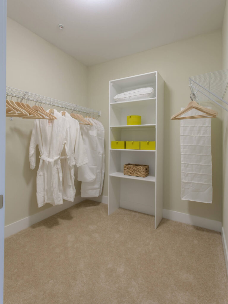 Walk-in closet with beige carpet, white walls, white shelving, yellow boxes, a wicker basket, folded linens, and several white robes hanging on wooden hangers.