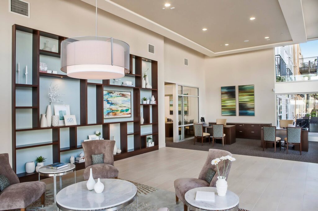 Modern office lobby with plush chairs, round marble tables, decorative shelves, abstract art, and a large ceiling light. Desks with chairs are in the background near large windows.