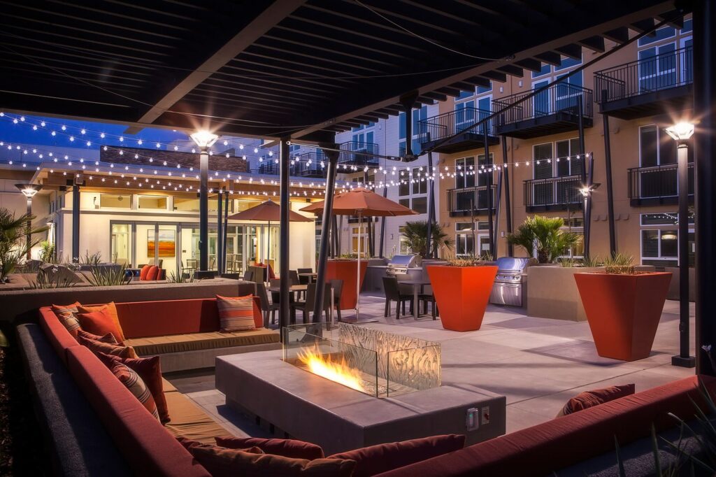 A modern outdoor patio at night with string lights, a rectangular fire pit, red seating, large orange planters, and grilling stations, surrounded by a multi-story apartment building with balconies.