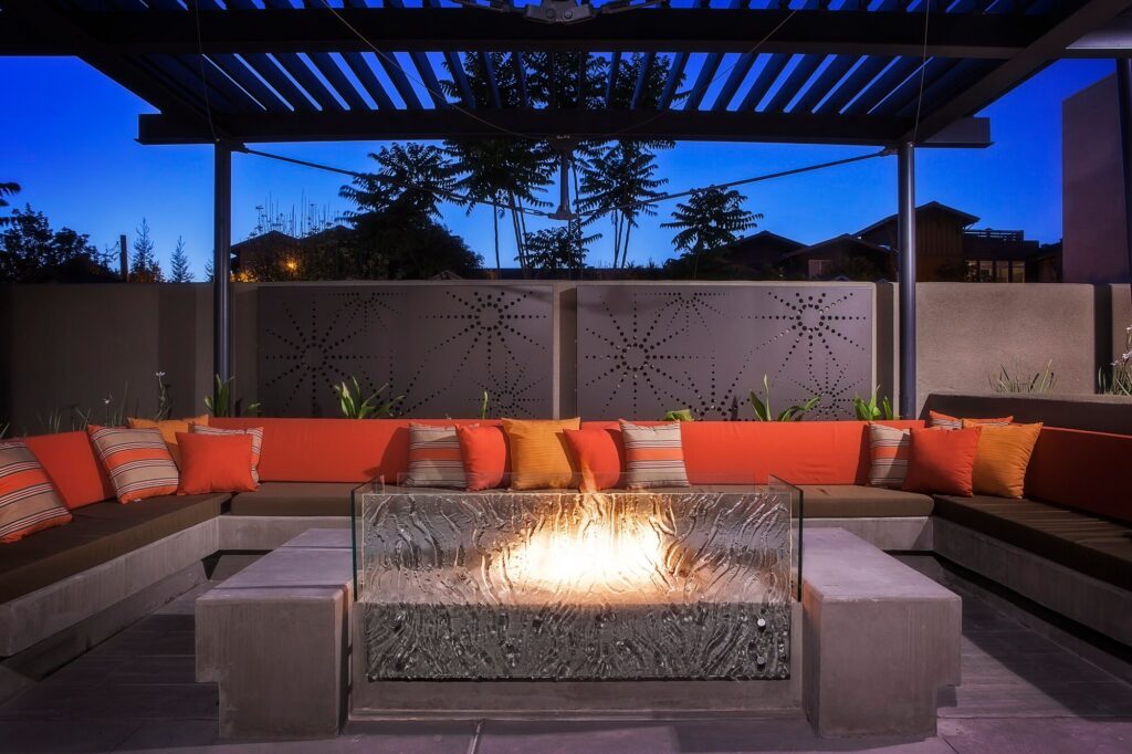 Modern outdoor patio at dusk with a U-shaped red cushioned sofa, colorful pillows, glass fire pit in the center, patterned privacy screen, and pergola overhead.