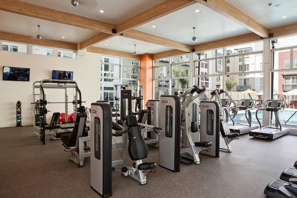 Modern gym with various weight machines, free weights, and cardio equipment in a bright room with large windows. A pool and apartment buildings are visible outside through the windows.