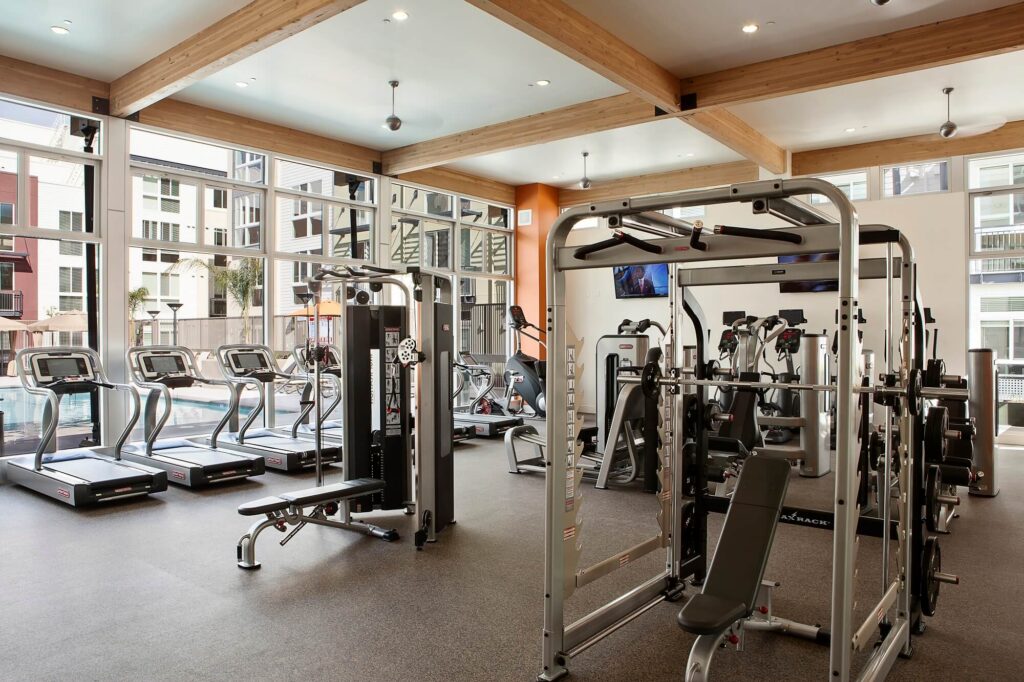 Modern gym with treadmills, weight machines, and exercise bikes. Large windows let in natural light, and there are apartment buildings visible outside. The gym has a spacious, clean layout with wooden beams on the ceiling.