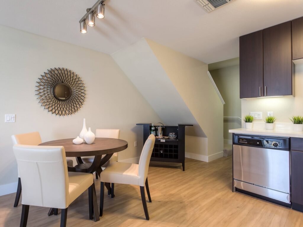 Modern dining area with a round table, four white chairs, a sunburst mirror on the wall, a small black cabinet, and a kitchen with a dishwasher and decorative plants on a light wood floor.