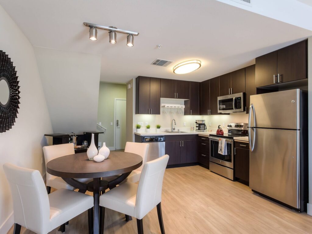 Modern kitchen with dark cabinets, stainless steel appliances, and a round dining table with four white chairs. Decorative vases sit on the table, and wall art hangs to the left. Light wood flooring throughout.