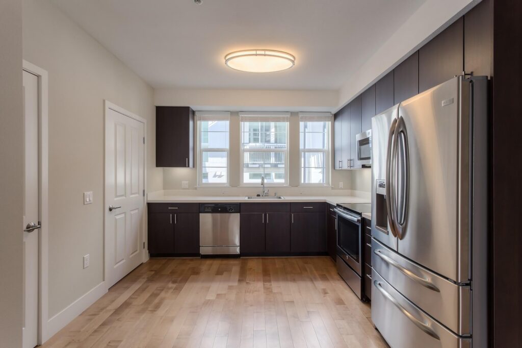 Modern kitchen with stainless steel appliances, dark cabinets, light wood floor, double-door fridge, oven, dishwasher, and a window above the sink letting in natural light.