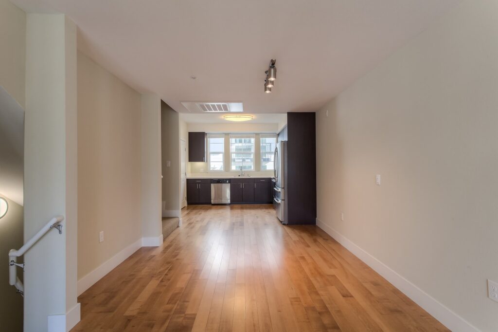 Empty apartment with light wood floors, beige walls, and a modern kitchen area in the back. Large windows provide natural light, and there are ceiling lights and a staircase to the left.