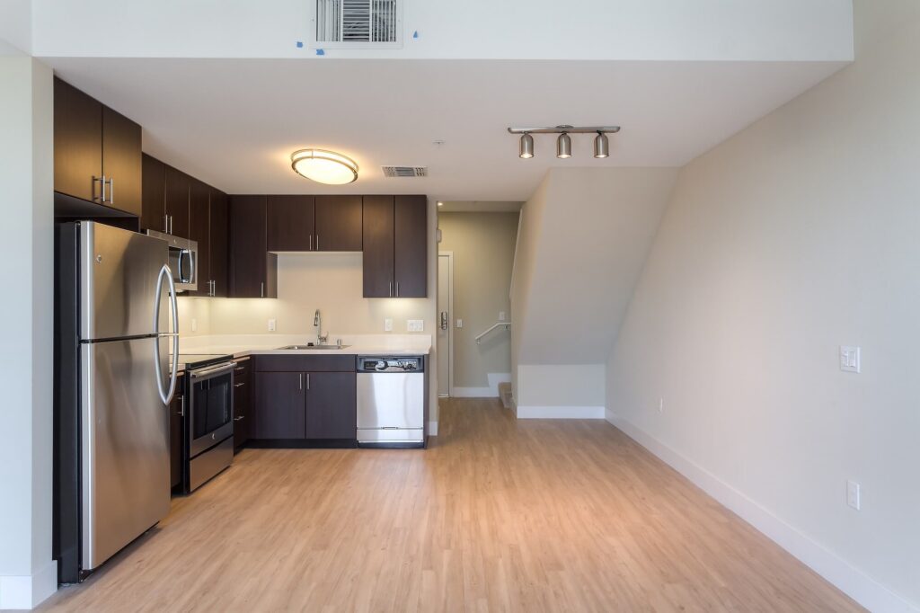 Modern kitchen with dark cabinets, stainless steel appliances, light wood flooring, and white walls. An open area leads to a stairway in the background. Ceiling lights brighten the space.