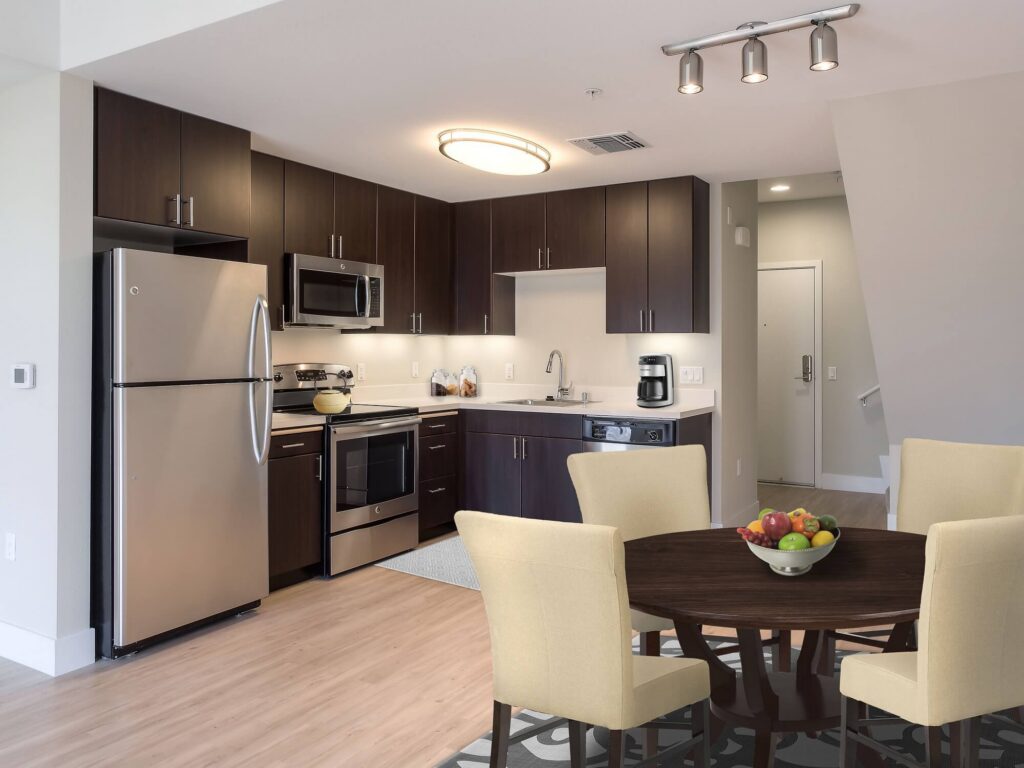 Modern kitchen with stainless steel appliances, dark wood cabinets, and a round dining table with four yellow chairs. A bowl of fruit sits on the table; a coffee maker is on the counter. Stairs and a white door are visible.