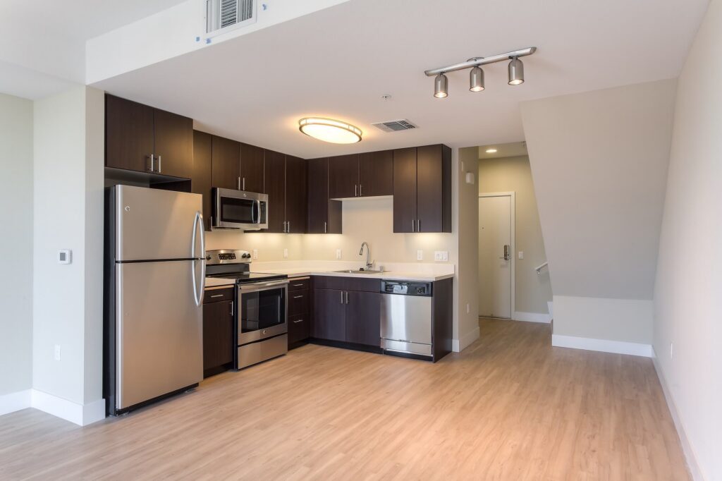 Modern apartment kitchen with stainless steel appliances, dark wood cabinets, light wood flooring, and white walls. Stairs and an entryway are visible to the right. Ceiling lights provide bright illumination.