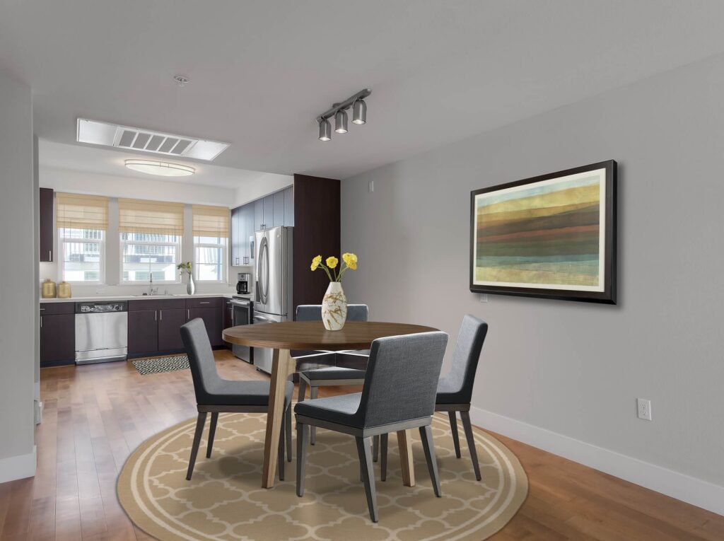 Modern dining area with a round table, four gray chairs, a vase with yellow flowers, wall art, hardwood floors, and an open kitchen in the background.