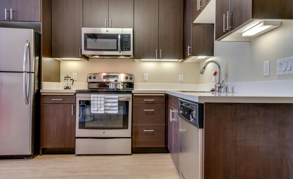 Modern kitchen with dark wood cabinets, stainless steel appliances including a fridge, oven, microwave, and dishwasher, under-cabinet lighting, and a white countertop with a sink and faucet.