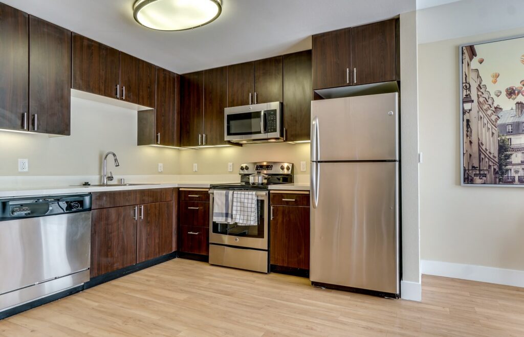 Modern kitchen with dark wood cabinets, stainless steel appliances, a dishwasher, stove, microwave, and refrigerator. Light wood flooring, white countertops, and a wall art piece on the right side. Ceiling light overhead.