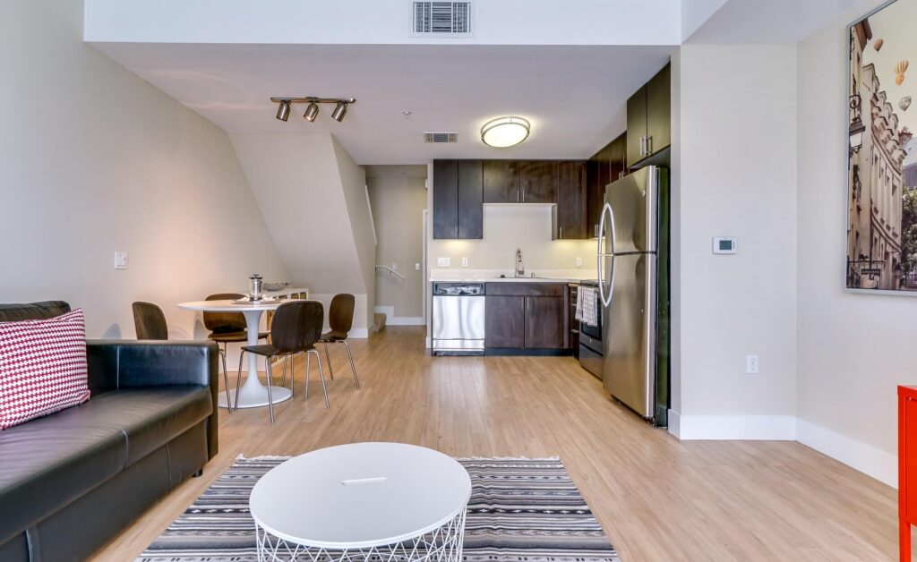 Modern open-plan apartment with a kitchen, dining area, and living room. Features wood flooring, stainless steel appliances, a round dining table, black couch, and a coffee table on a striped rug.