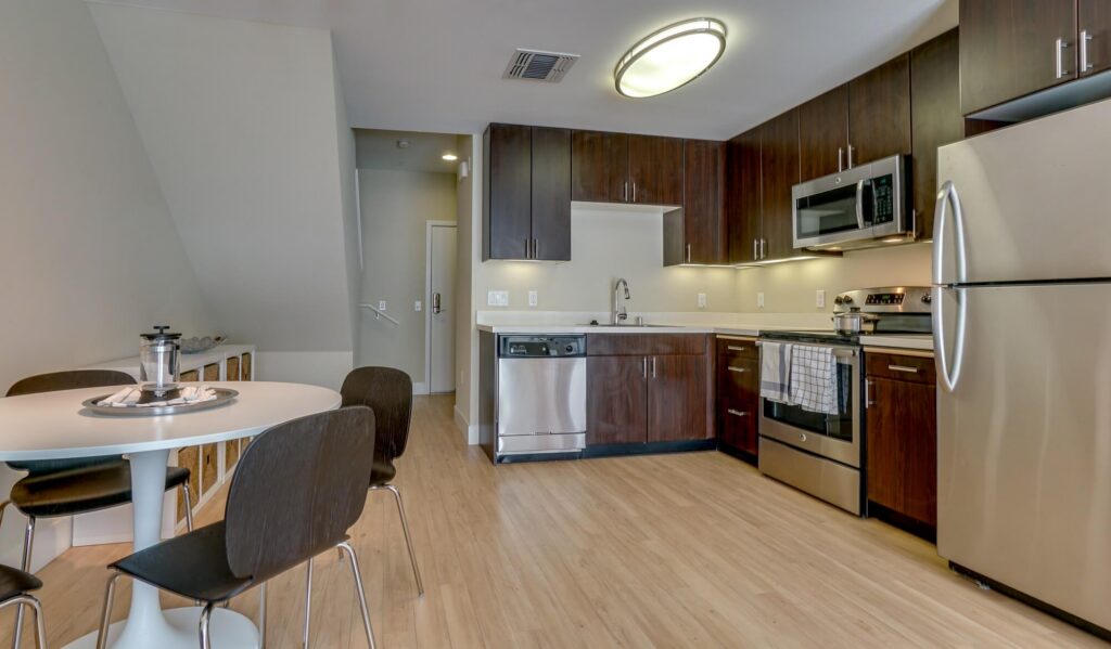 Modern kitchen with stainless steel appliances, dark wood cabinets, and a small dining table with four chairs on light wood flooring.