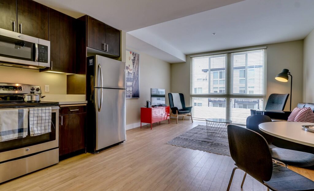Modern apartment kitchen and living area with stainless steel appliances, dining table, chairs, armchairs, TV, and large window letting in natural light.