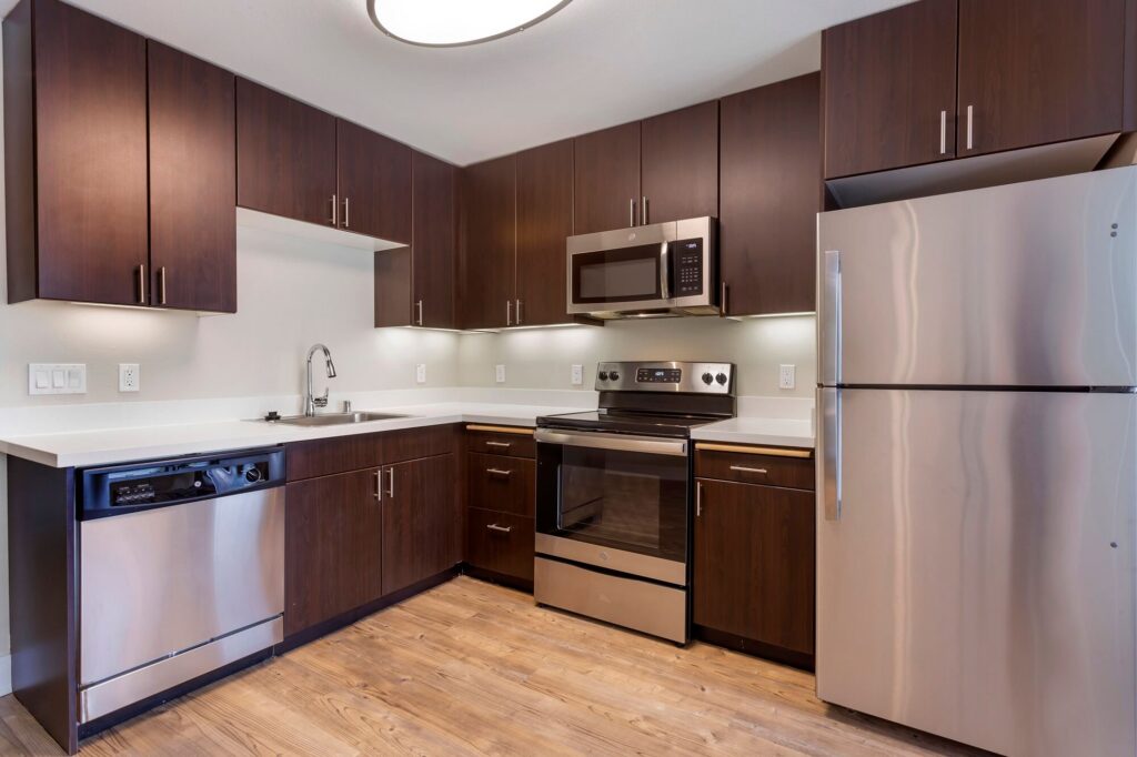 Modern kitchen with dark wood cabinets, stainless steel appliances including refrigerator, stove, microwave, and dishwasher, white countertops, and light wood flooring. Bright lighting and minimal decor.