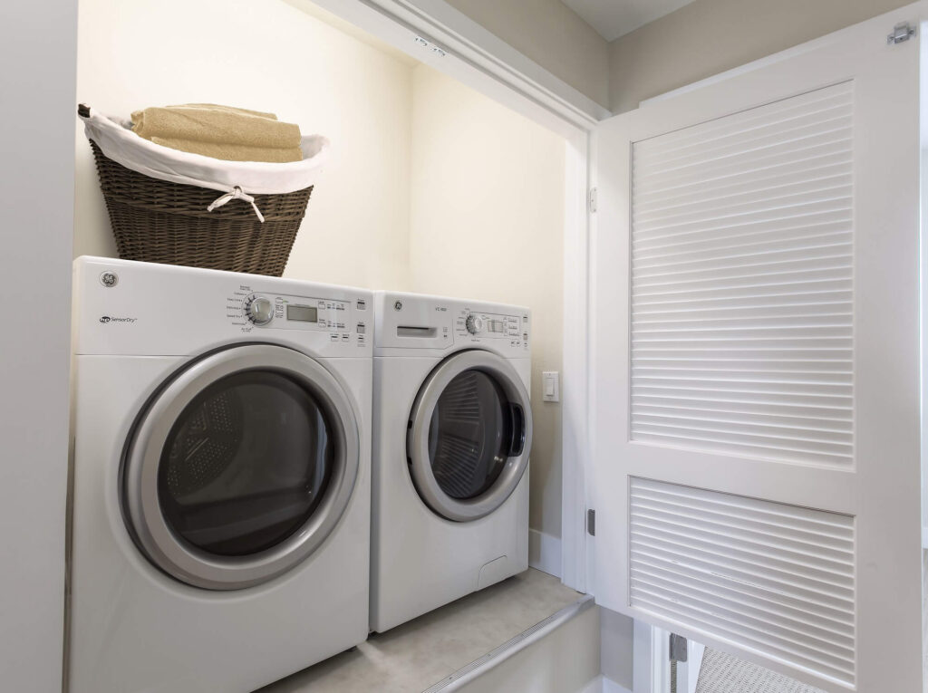 A modern laundry area with a white front-loading washer and dryer side by side. A dark woven basket with folded towels sits on top of the dryer. The space has white walls and a sliding white louvered door.