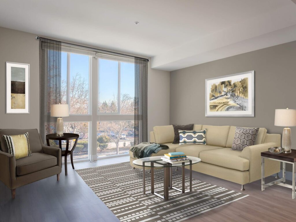 Modern living room with large windows, beige sofa, accent chair, coffee table, lamps, patterned rug, and two landscape artworks on the walls. Natural light fills the space, highlighting neutral colors and cozy decor.