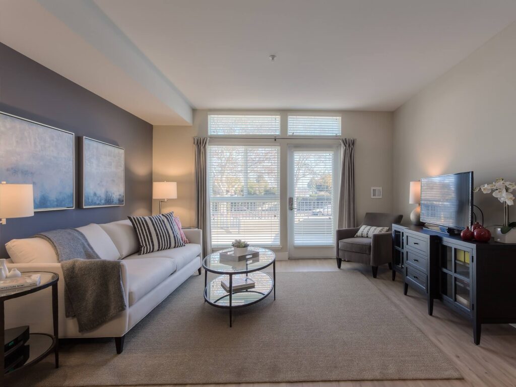 Modern living room with a white sofa, armchair, TV, coffee table, and art on the wall. Large windows let in natural light, creating a cozy and bright atmosphere.