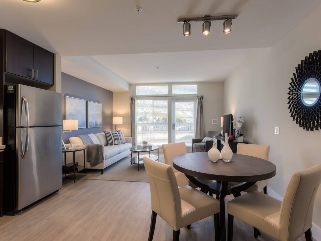 Modern apartment interior with a round dining table, four beige chairs, a sofa, coffee table, TV, wall art, large windows, and stainless steel refrigerator. Warm lighting brightens the neutral-toned space.