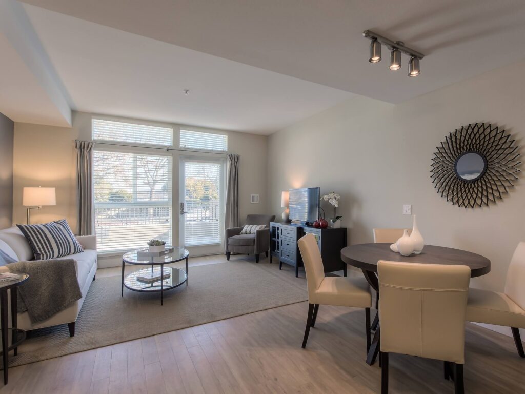 Modern living room with a sofa, round coffee table, TV on a console, dining table with four chairs, large windows with curtains, and neutral-colored decor.