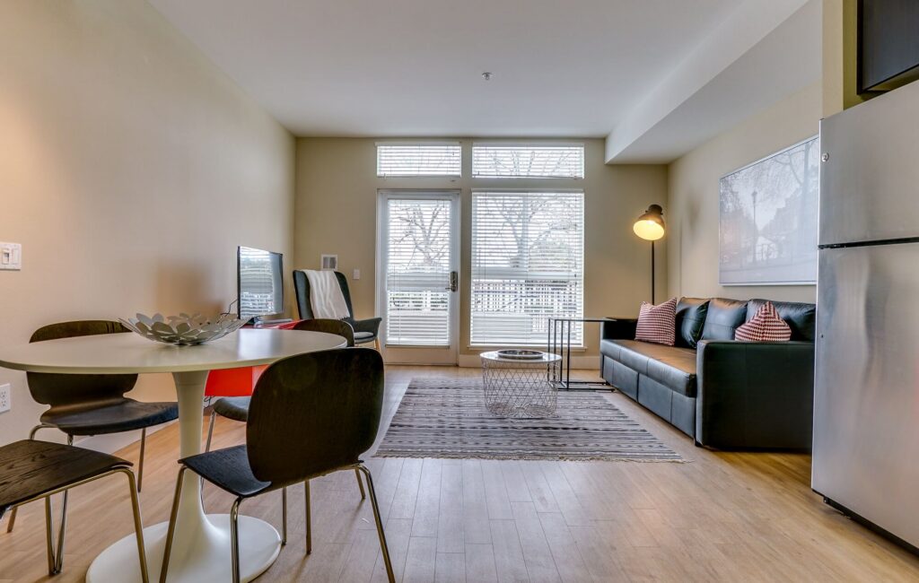 Modern living room and dining area with a round table and four chairs, a black sofa with striped pillows, a coffee table, large windows, and a stainless-steel refrigerator on wood flooring.