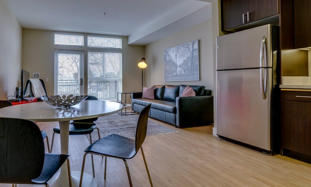 Modern apartment with wood floors, round dining table, four chairs, stainless steel fridge, dark cabinets, sofa with pillows, floor lamp, TV, and large windows letting in natural light.