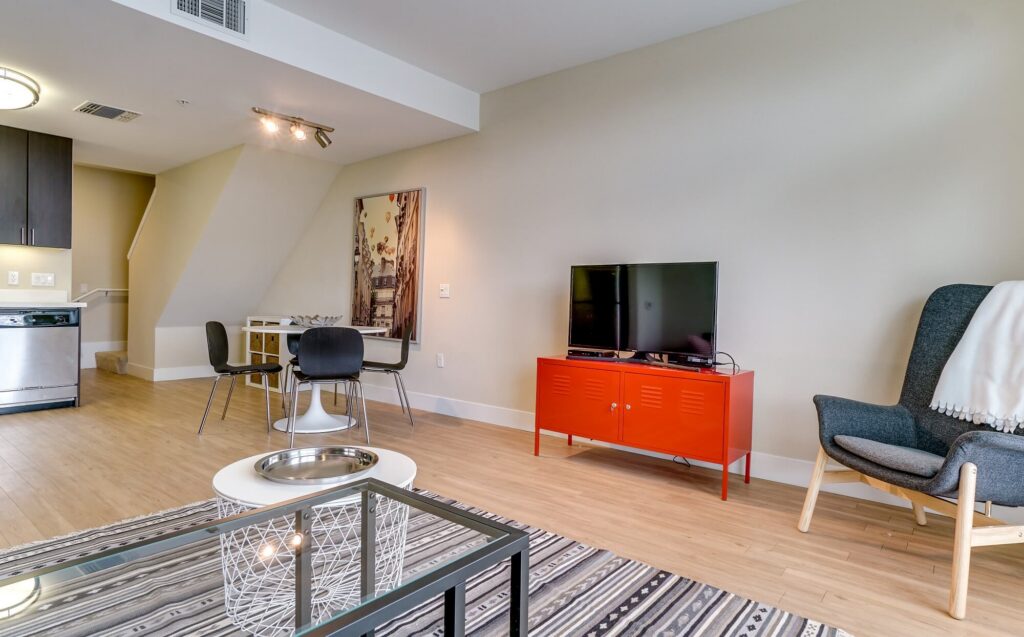 Modern living room with a TV on a red cabinet, gray armchair with a blanket, small dining table with two chairs, and a striped rug on a light wood floor. Kitchen area visible in the background.