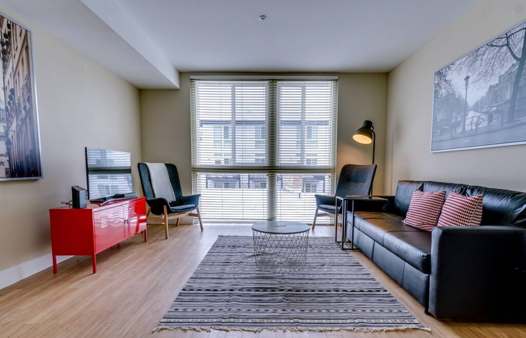 Modern living room with a black leather sofa, striped rug, coffee table, TV on a red stand, two chairs, floor lamp, and large window with blinds, letting in natural light. Framed art decorates the beige walls.